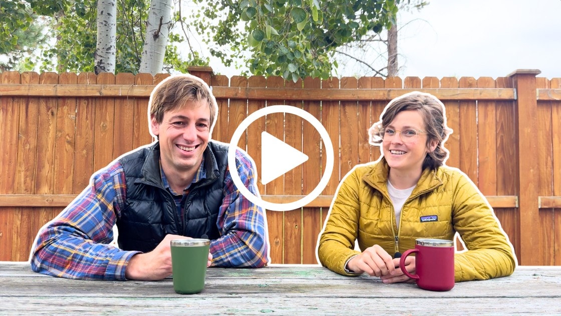 Two people sit at an outdoor wooden picnic table with mugs; one wears a plaid shirt and vest, the other a yellow jacket.