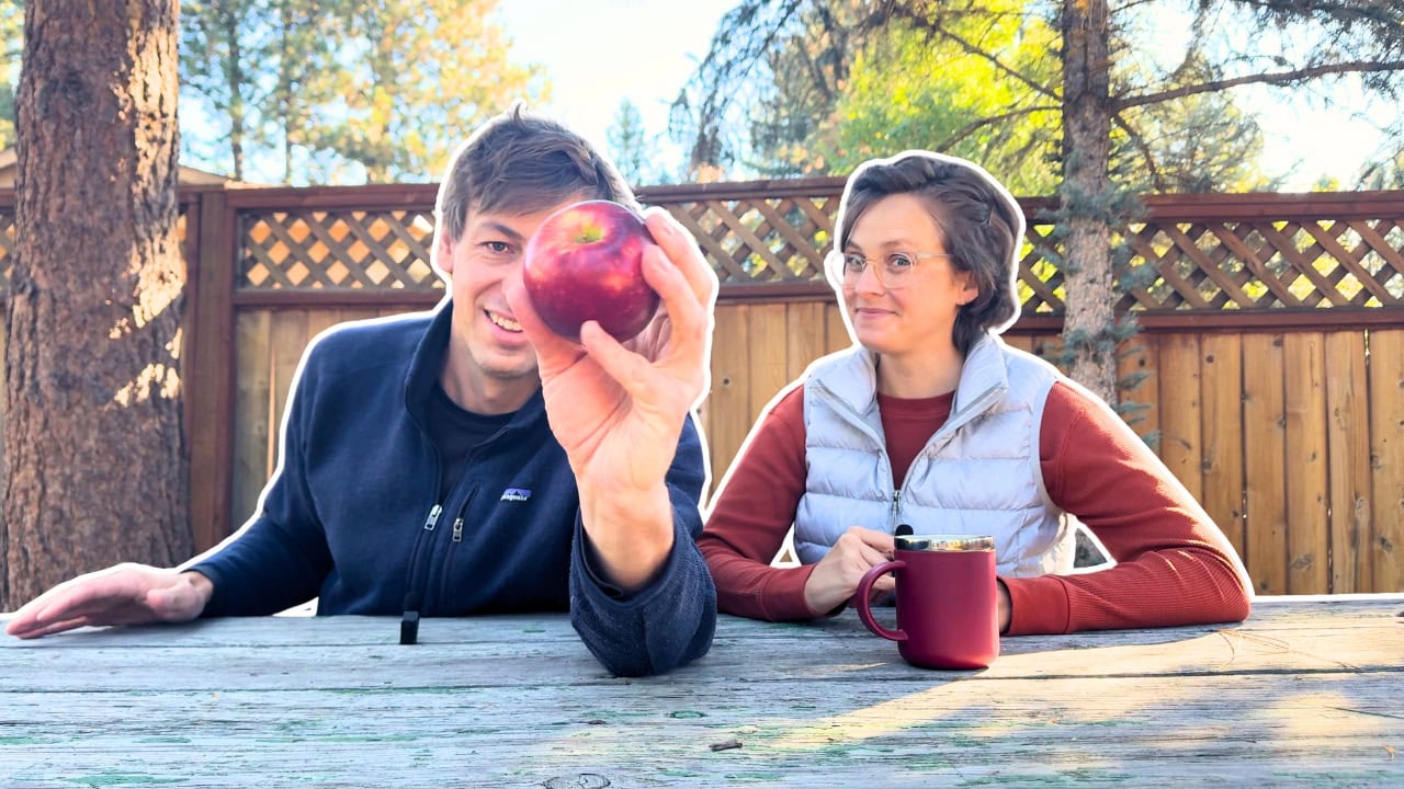 Two people sit at an outdoor wooden table; one holds a red apple, the other, in glasses and white vest, holds a mug. Trees and fence behind.