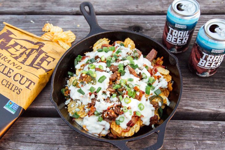 Nachos with steak and beer-cheese sauce in a cast iron skillet. Bag of kettle chips and can of beer in frame.