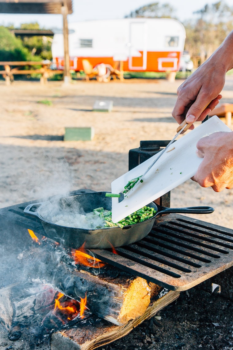 Michael transferring chopped asparagus from a cutting board to a cast iron skillet on a campfire