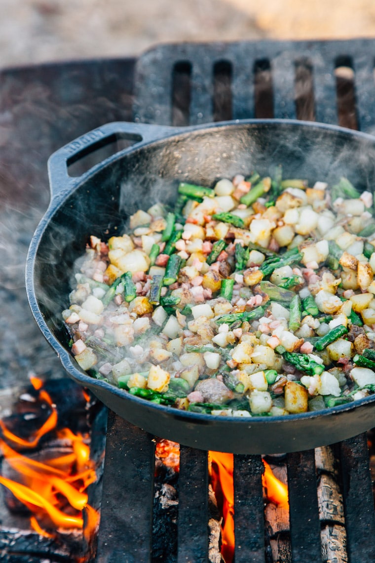 Potato hash and asparagus cooking in a cast iron skillet over a campfire