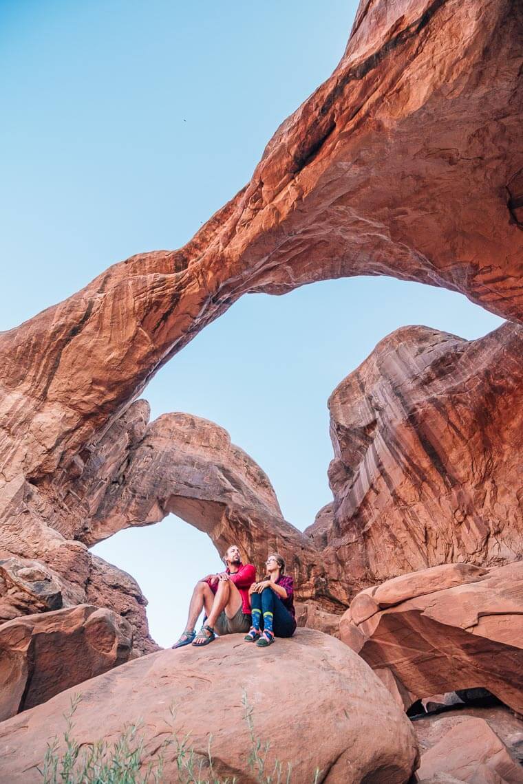 Megan and Michael sitting on a rock looking up at the double arch in Arches national Park