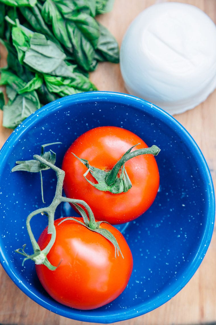 Two ripe tomatoes in a bowl next to fresh basil and mozzarella
