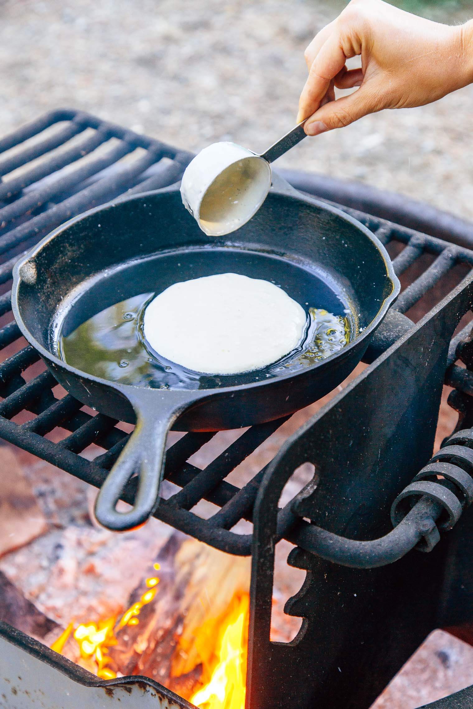 Megan scooping pancake batter into a cast-iron skillet that is over a campfire