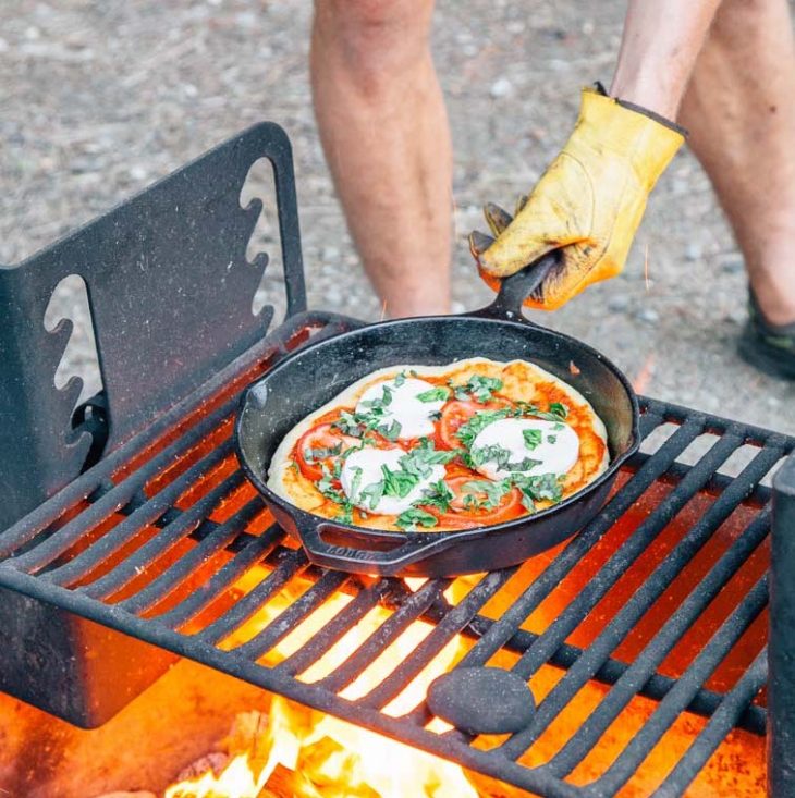 Michael picking up a cast iron skillet filled with pizza off of a campfire grill