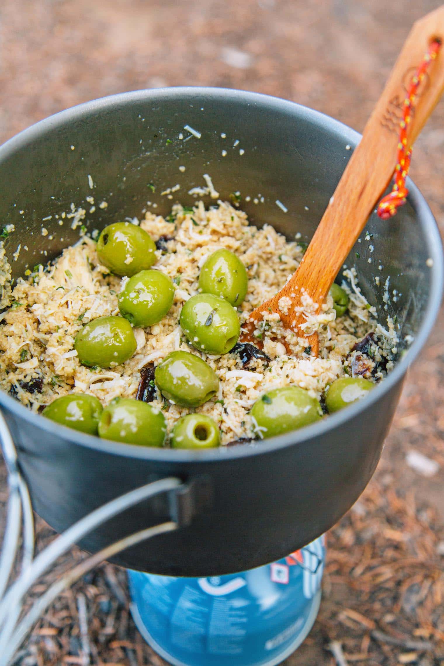 A wooden spoon in a pot that is filled with couscous and green olives