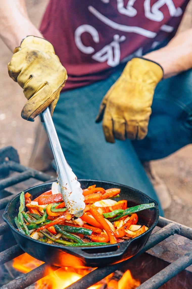 Michael using tongs to toss sliced peppers and onions that are cooking in a cast iron skillet over a campfire