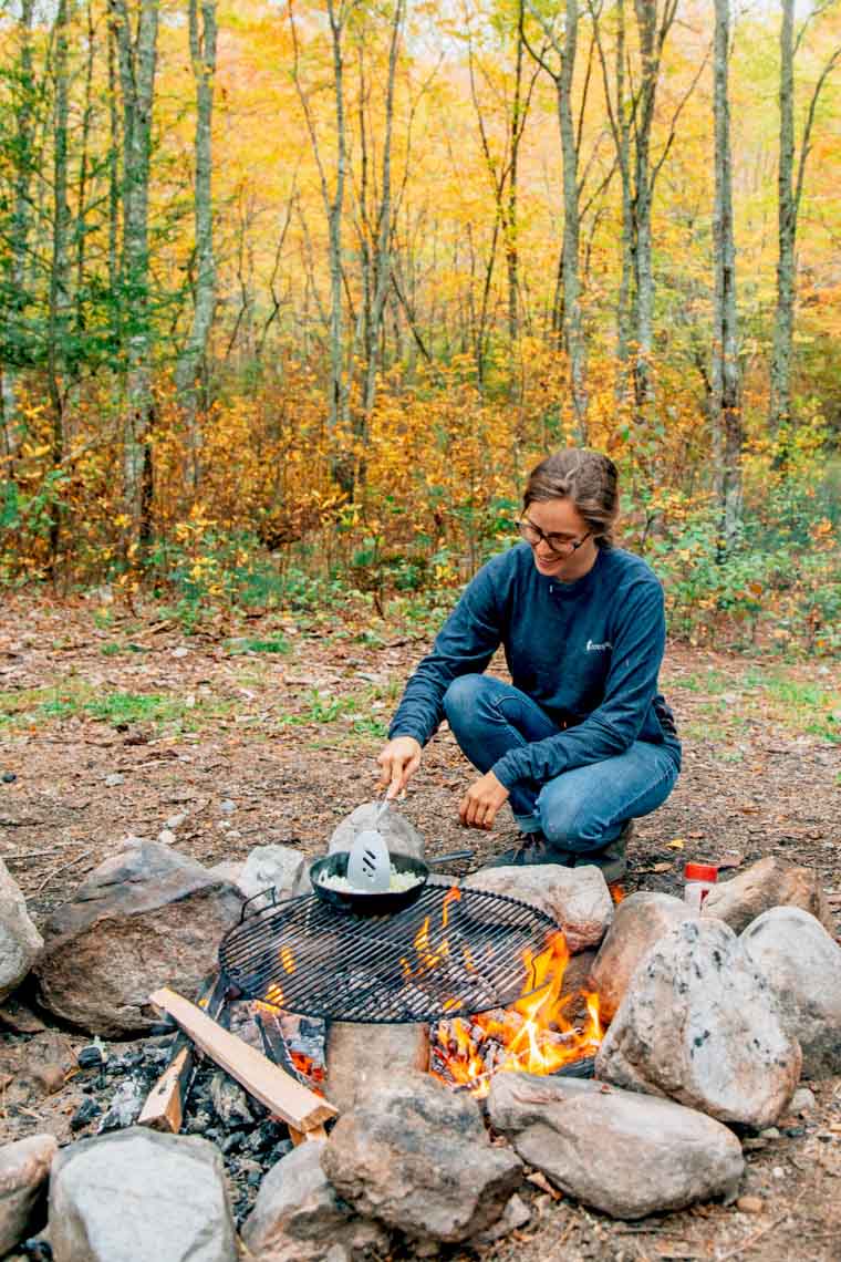 Megan cooking over a campfire with fall foliage in the background