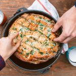 Two hands picking up  pieces of pull apart garlic bread in a Dutch oven
