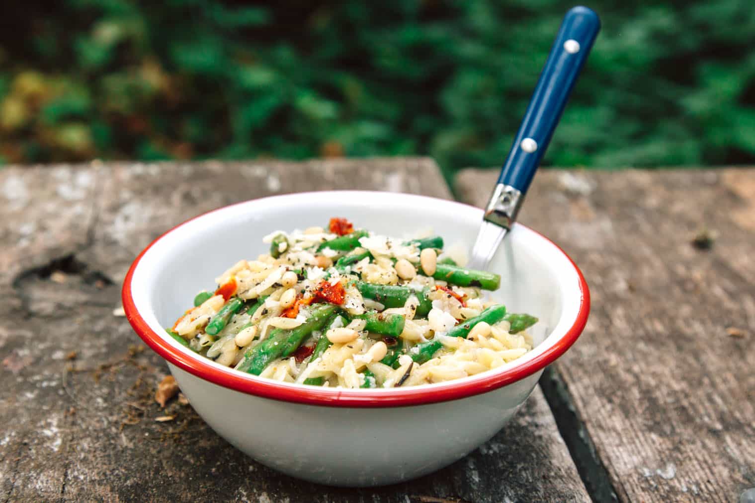 A white enamel bowl that is filled with asparagus and pasta on a camping table