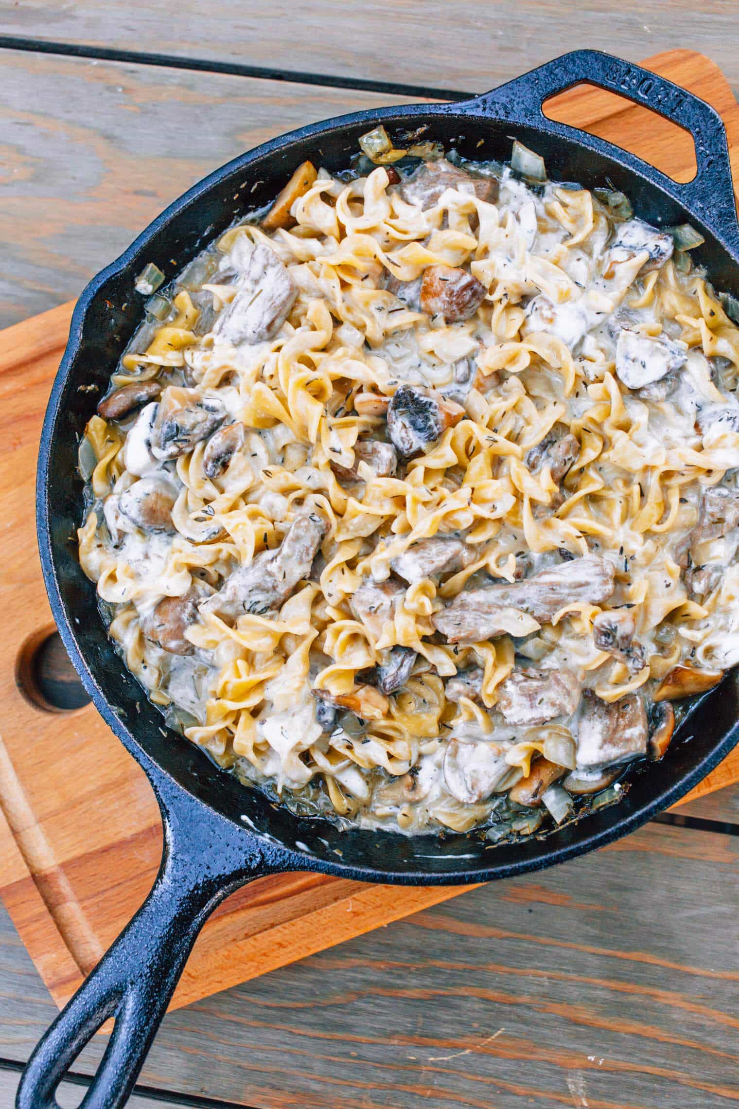 A cast-iron skillet filled with beef stroganoff resting on a wooden cutting board