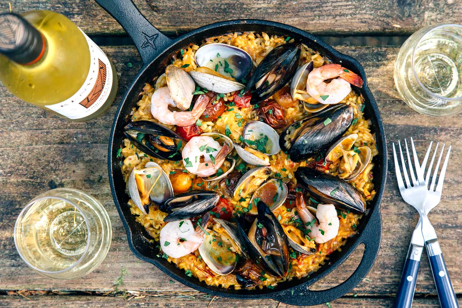 Paella in a skillet on a camping table with forks and a bottle of wine in frame