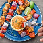 Thai grilled chicken skewers on a blue camping plate with a small bowl of peanut dipping sauce.