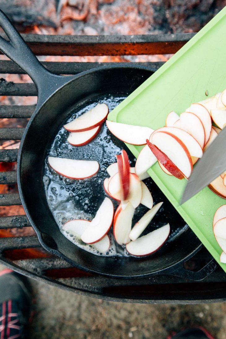 Moving sliced apples from a cutting board into a skillet over a campfire