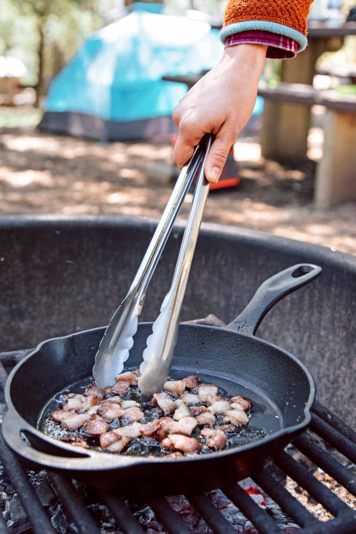 Megan using tongs to cook chopped bacon in a cast iron skillet over a campfire