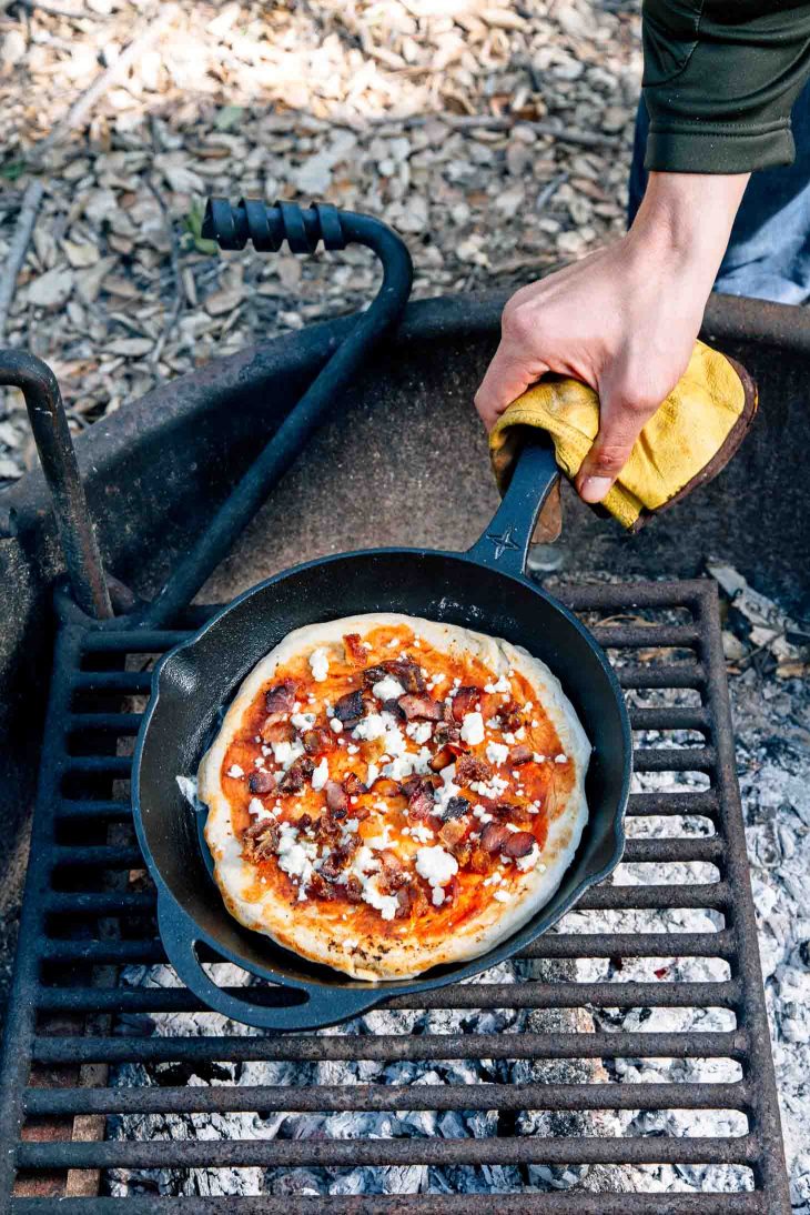 Michaels gloved hand lifting a cast iron skillet of pizza off of the campfire
