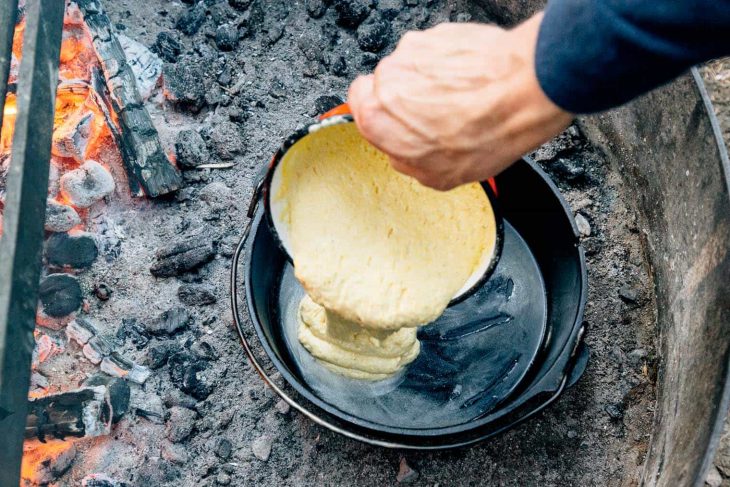 Michael pouring cornbread batter into Dutch oven in a fire pit