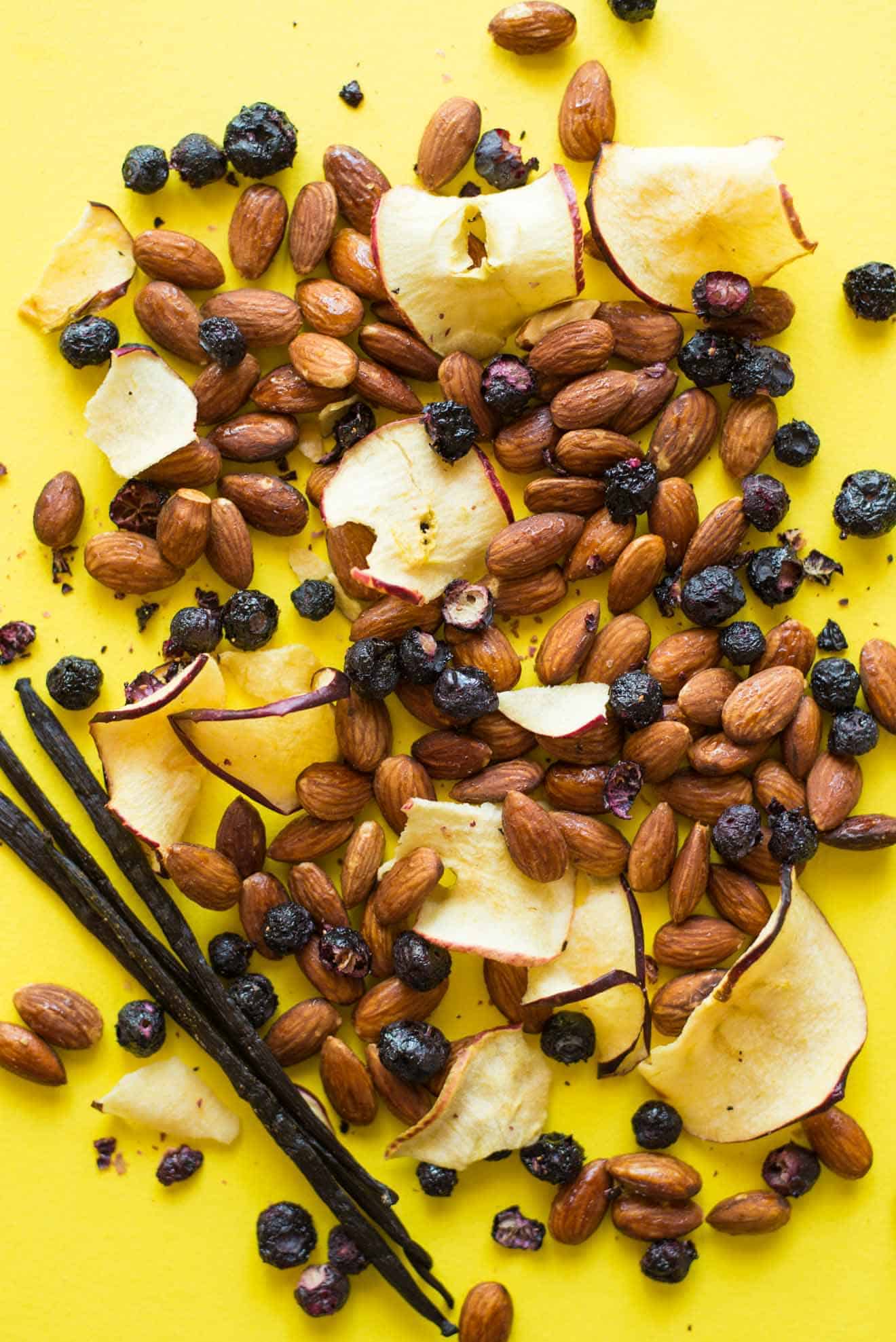 Dried apple slices, almonds, raisins and blueberries are spread on a bright yellow surface with a vanilla bean.