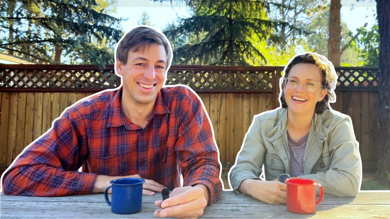 Two people sit at a wooden table outdoors with one blue mug and one red mug; tall trees and a wooden fence are in the background.