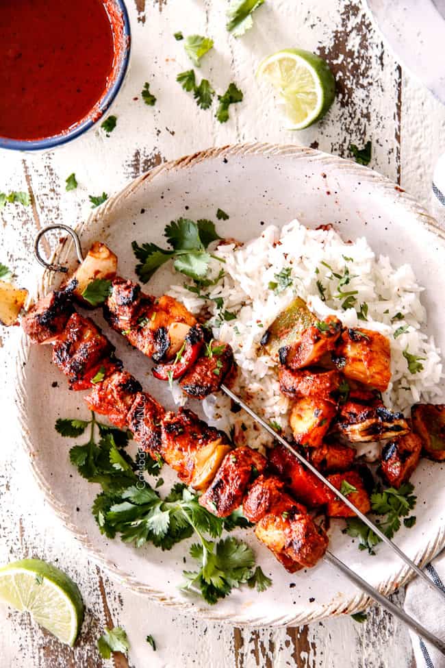 Grilled skewers of succulent chicken and vegetables served over a bed of fluffy rice, garnished with fresh cilantro and accompanied by a tangy dipping sauce and lime wedges, presented on a rustic ceramic plate.