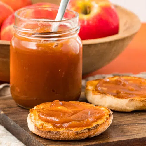 A jar of apple butter next to an english muffin topped with apple butter.