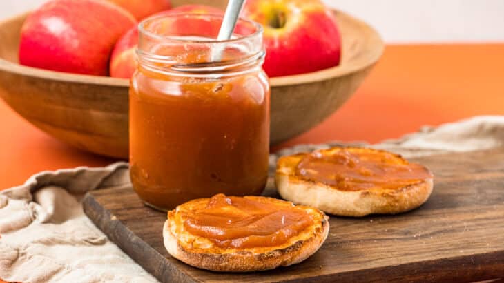 A jar of apple butter next to an english muffin topped with apple butter.