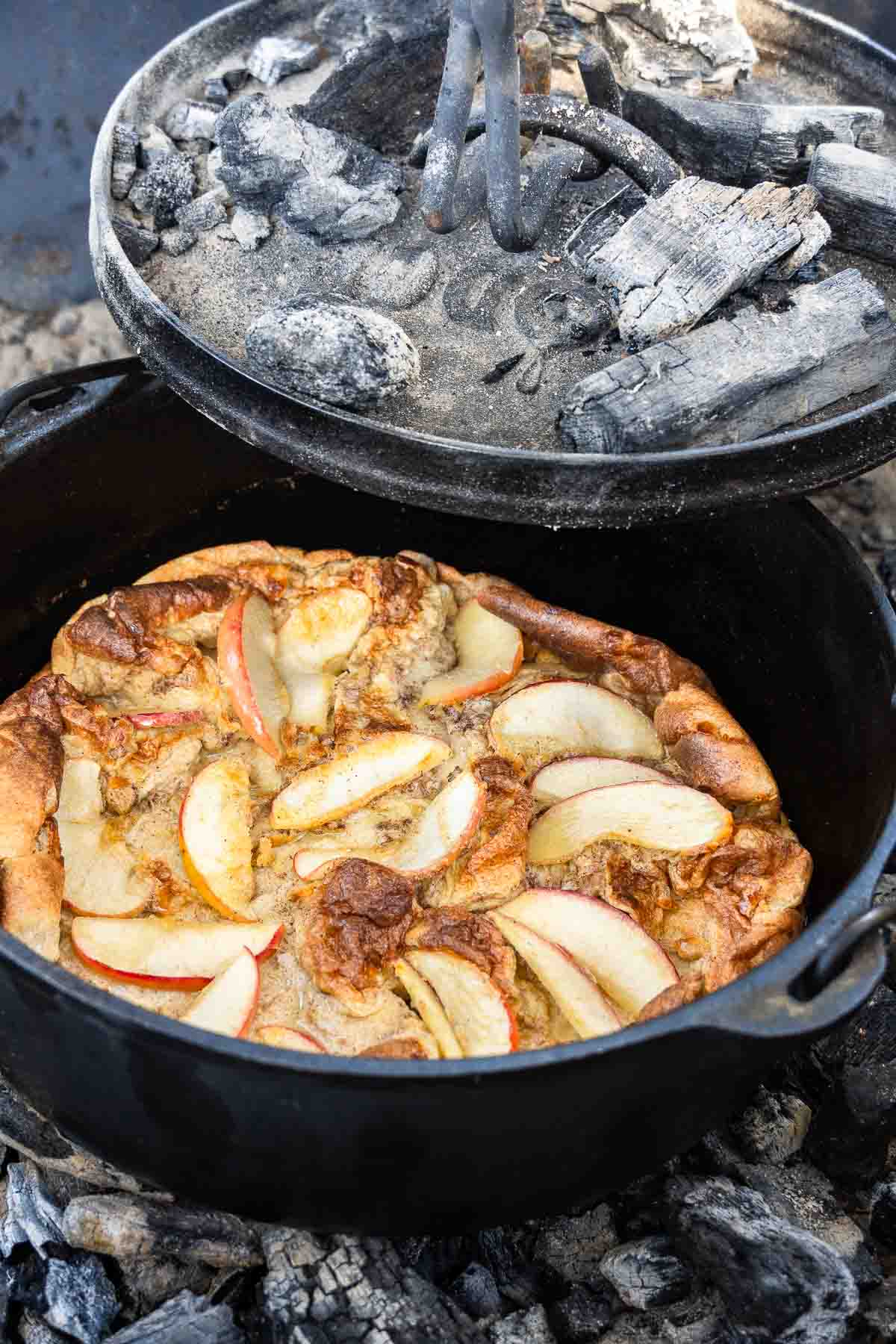 A Dutch oven with an apple dutch baby topped with apple slices sits on hot coals, lid partially open to show ash and charcoal.