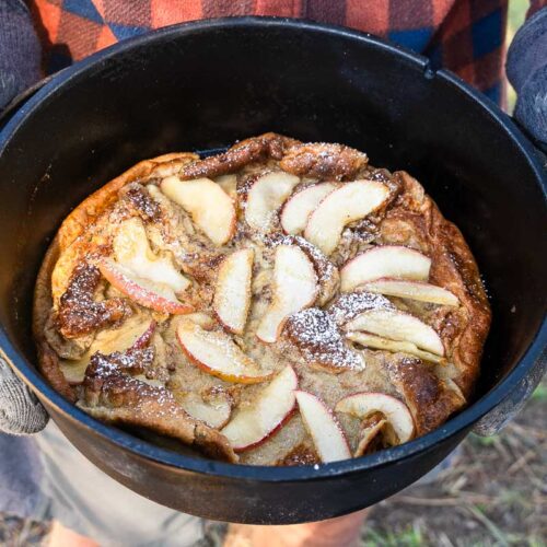 A person in gloves and a plaid shirt holds a cast iron pot with a dessert topped with apple slices and powdered sugar outdoors.