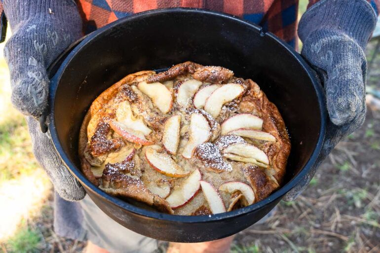 A person in gloves and a plaid shirt holds a cast iron pot with a dessert topped with apple slices and powdered sugar outdoors.