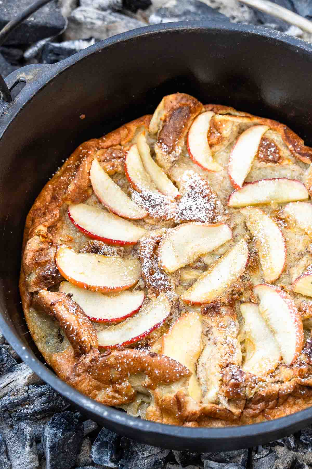 A baked apple pancake with sliced apples and powdered sugar sits in a black cast iron skillet over gray wood coals.