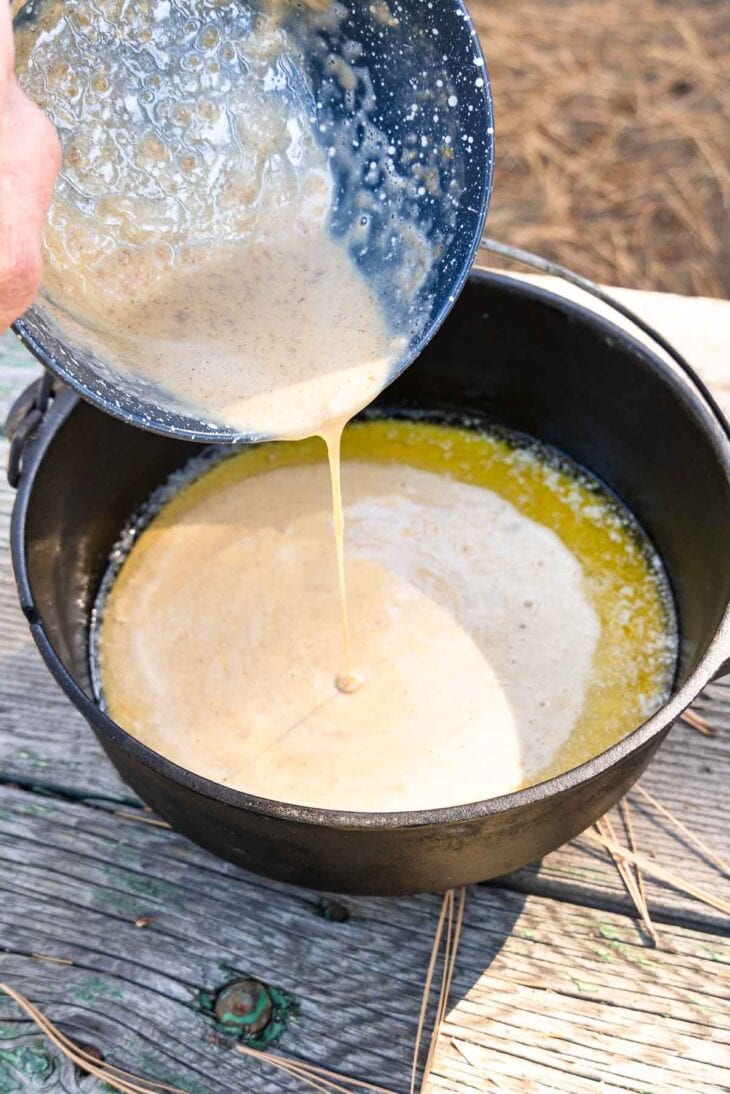 A person pours pale batter from a speckled blue bowl into a black cast iron pot with melted butter on a rustic wooden table outdoors.