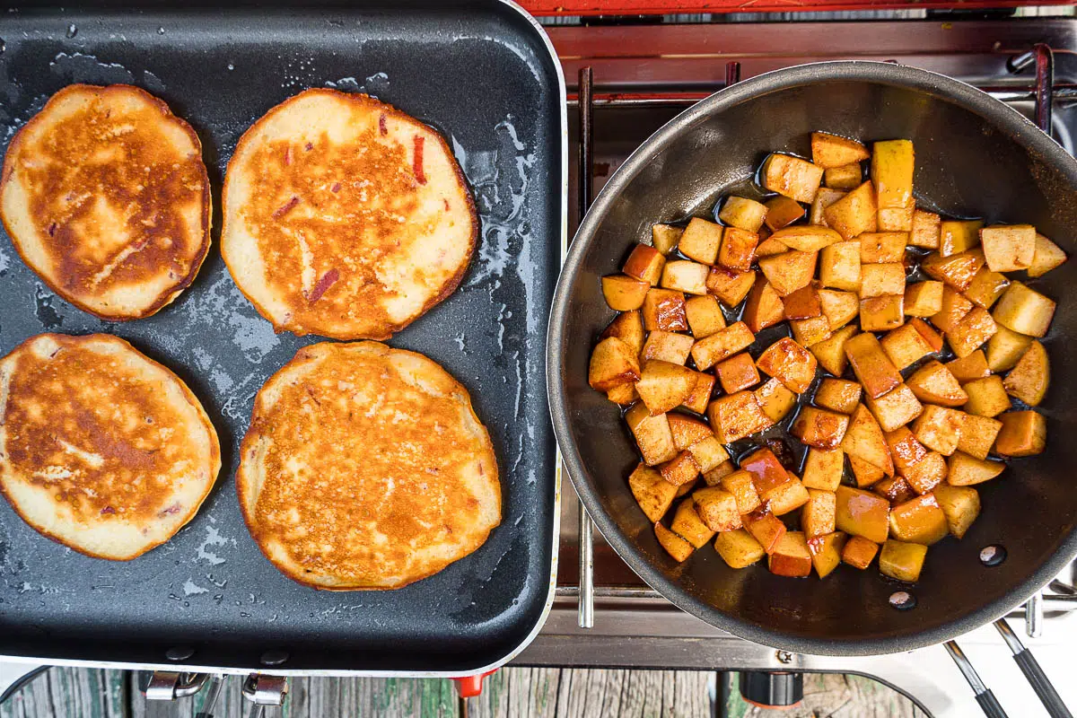 Four apple pancakes in a square skillet. Next to that is a round skillet with chopped apples.