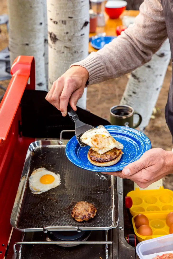 Michael is holding a plate with an english muffin and sausage on it. He is using a spatula to place an egg on top.