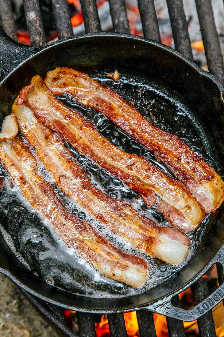 Four strands of bacon frying in a cast iron skillet over a campfire.