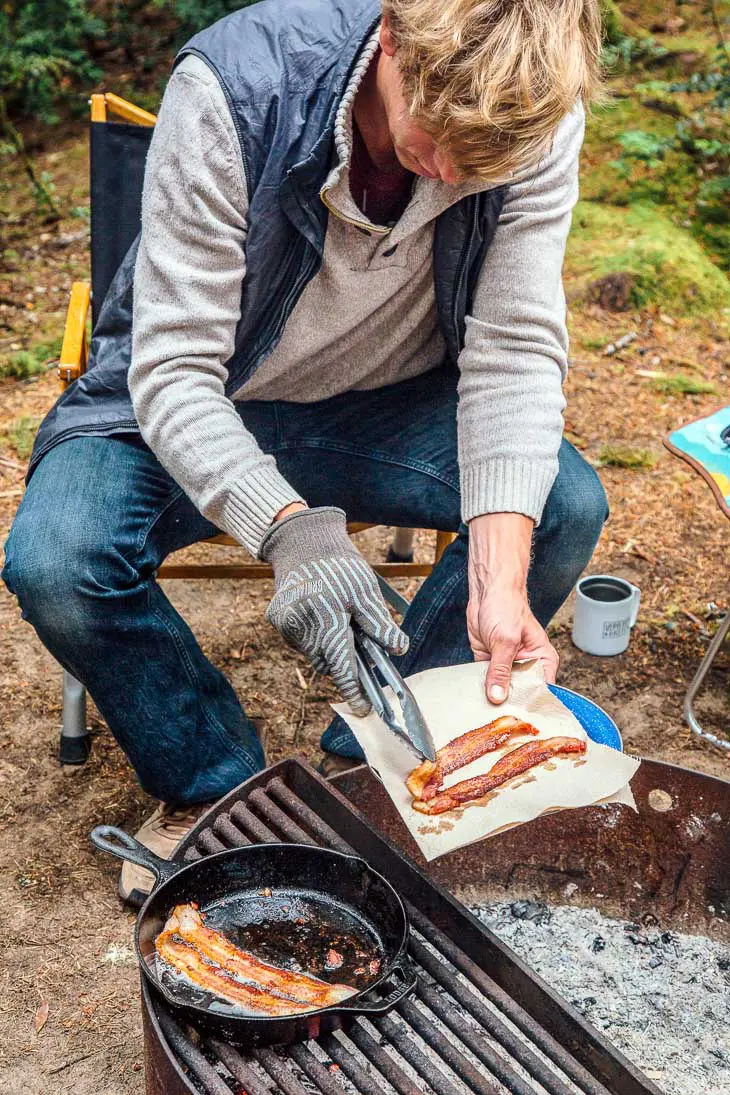 Michael is sitting by a campfire frying bacon in a cast iron skillet.