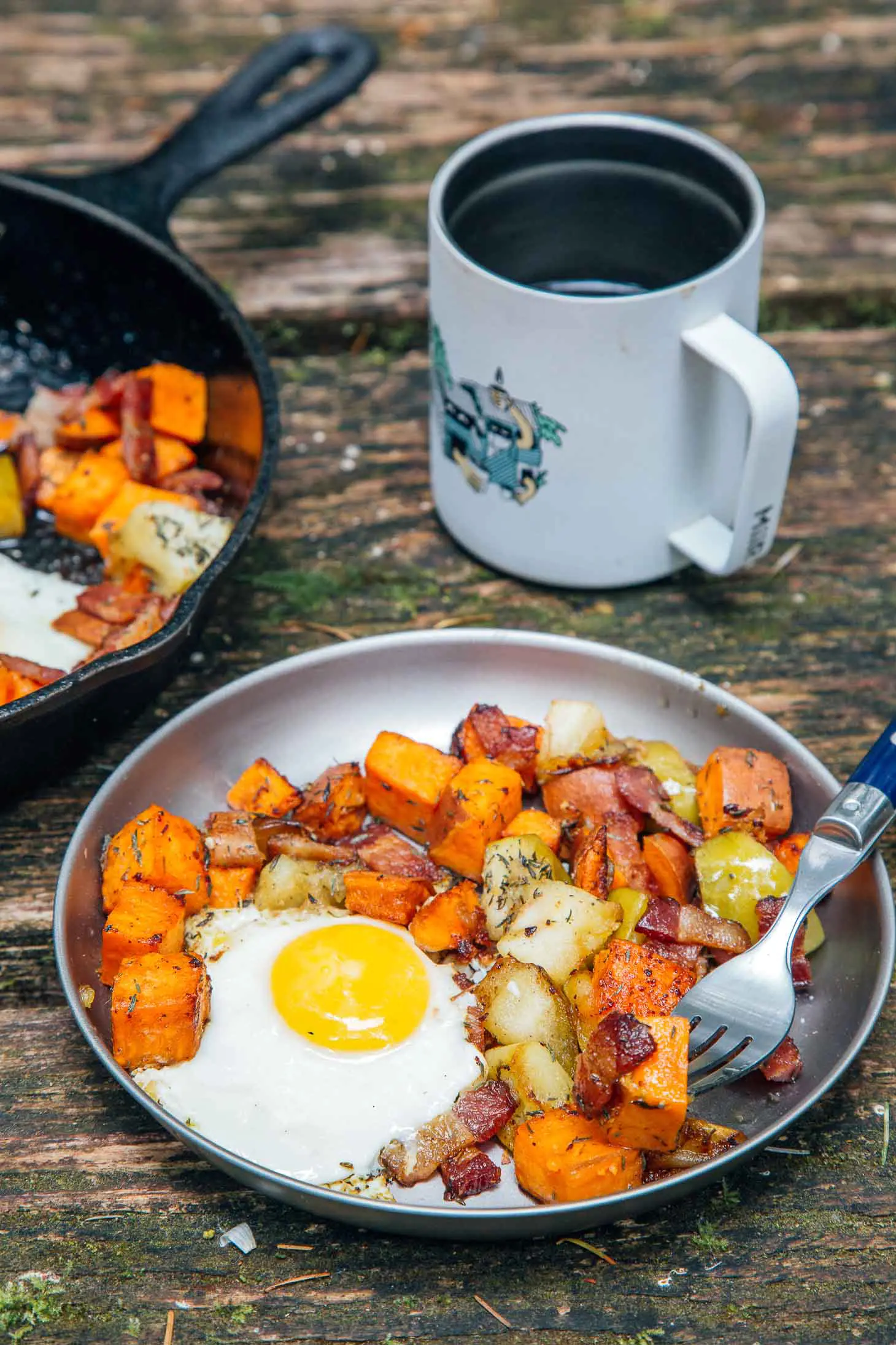 Apple and sweet potato hash & a fried egg on a silver plate on a camp table with a cup of coffee in the background.