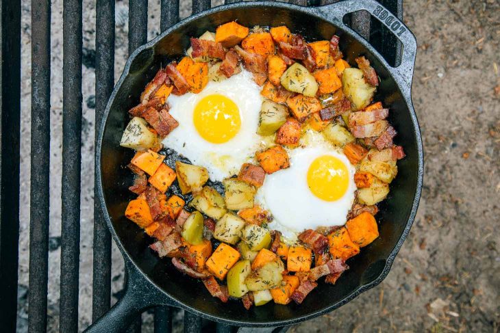 Overhead view of apple and sweet potato hash with two fried eggs in a cast iron skillet over a campfire.