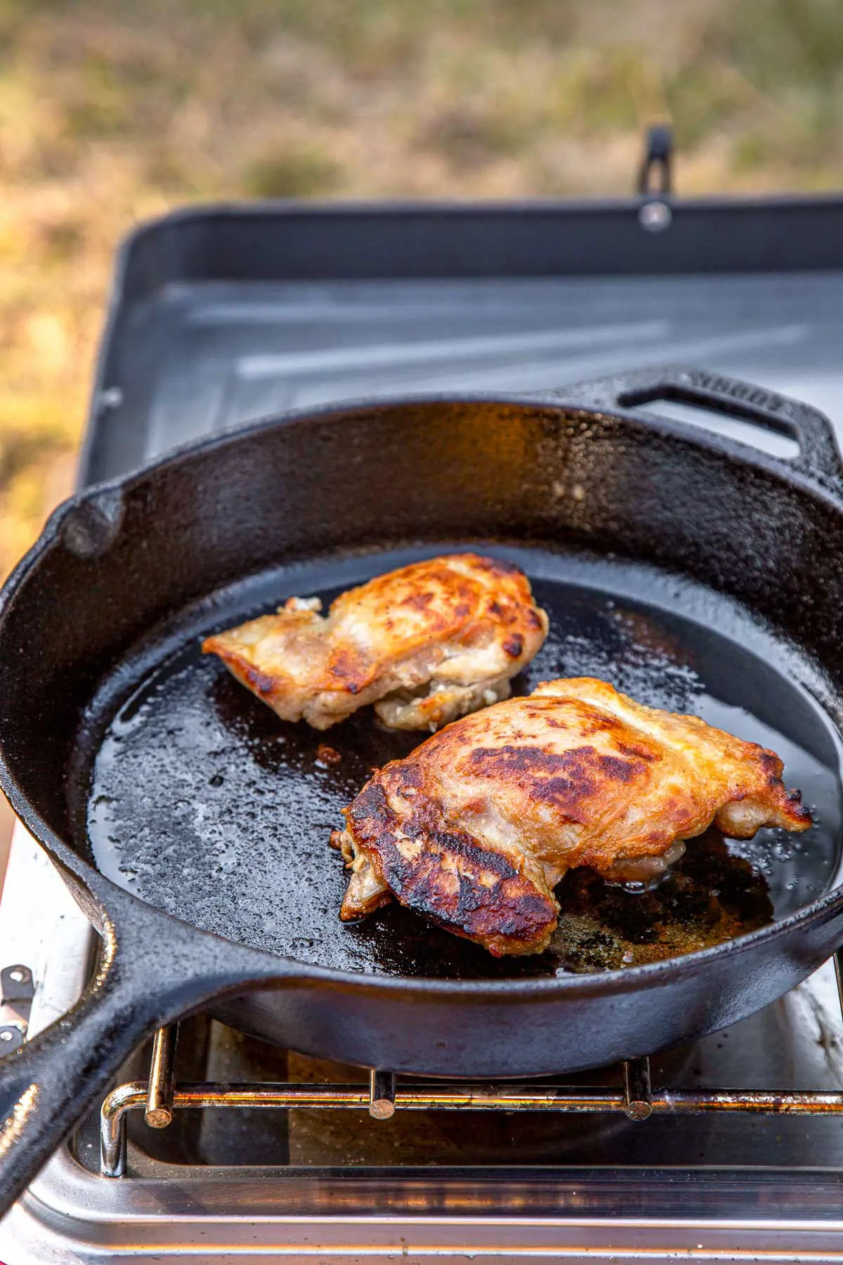 Two browned chicken thighs in a cast iron skillet on a camping stove.