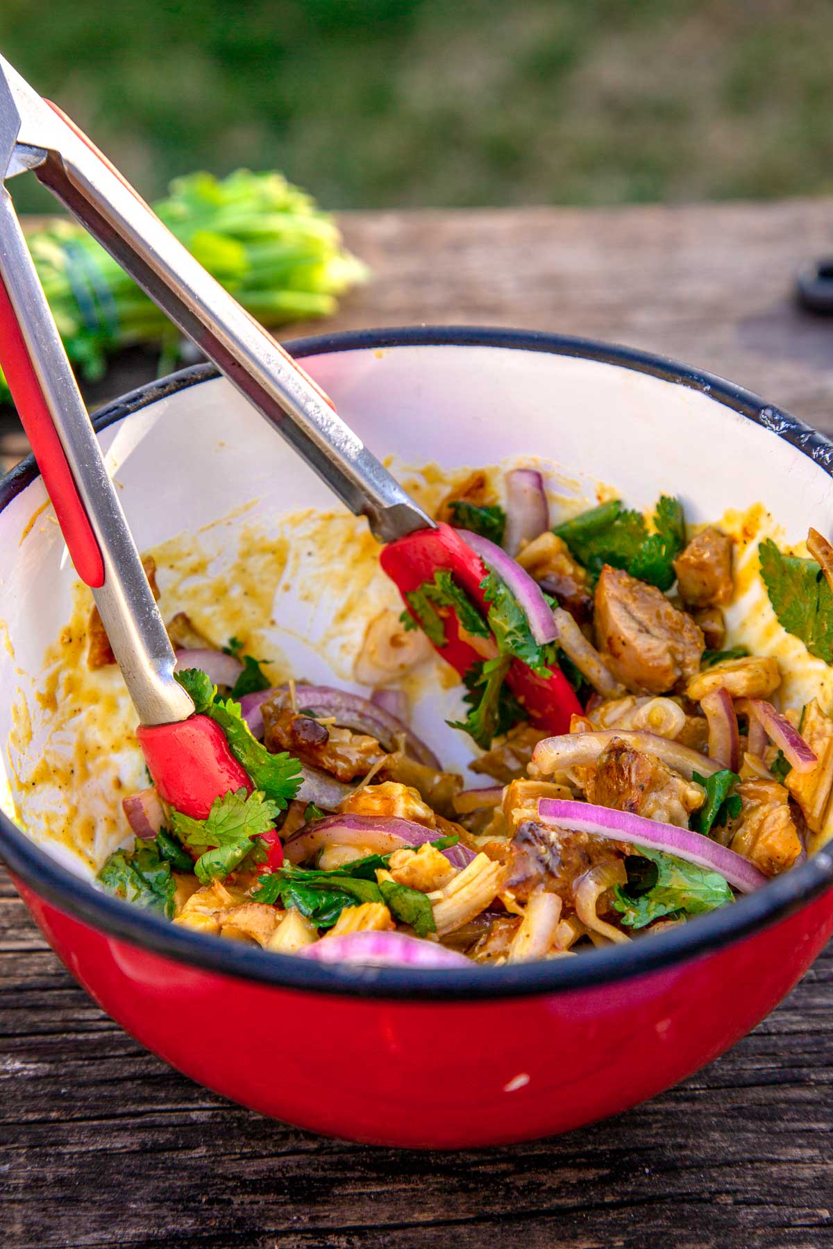 A red bowl full of BBQ sauced chicken, red onion, and cilantro. A pair of red tongs are resting in the bowl.