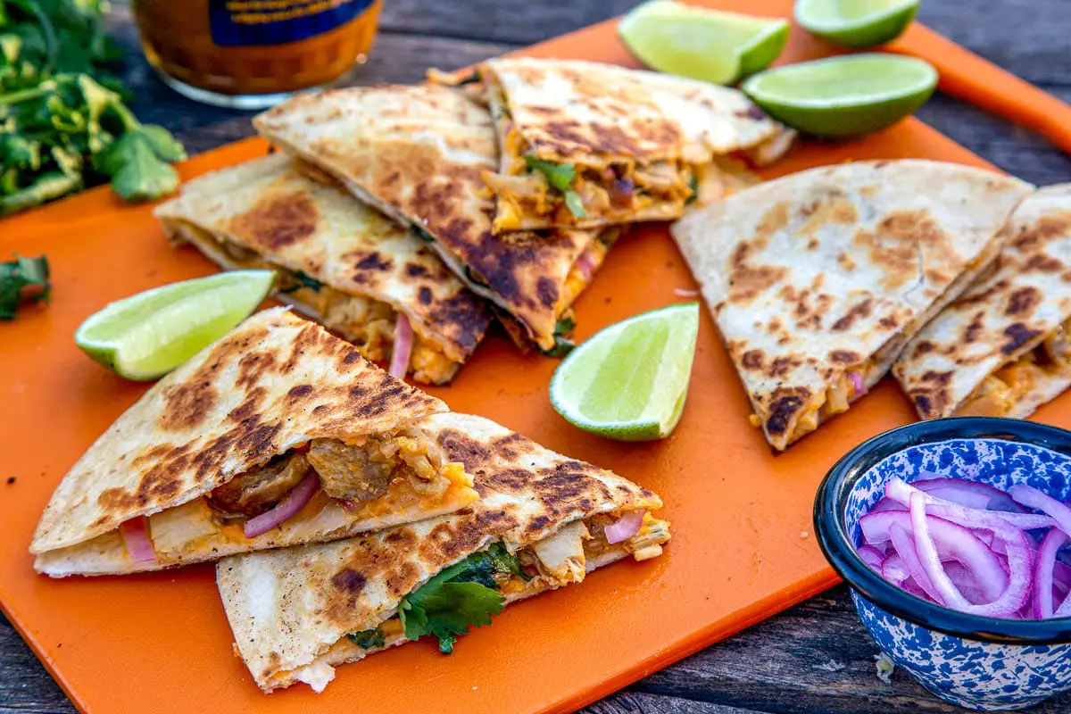 Sliced quesadillas stacked on an orange cutting board with slices of lime and a small bowl of red onions.