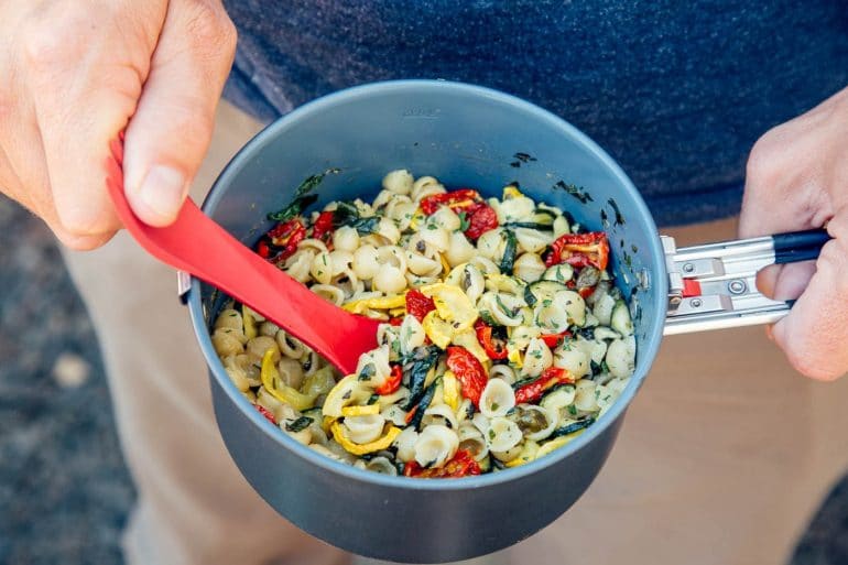 A person in a blue shirt and beige pants is holding a pot of cooked pasta with greens, tomatoes, and yellow squash.