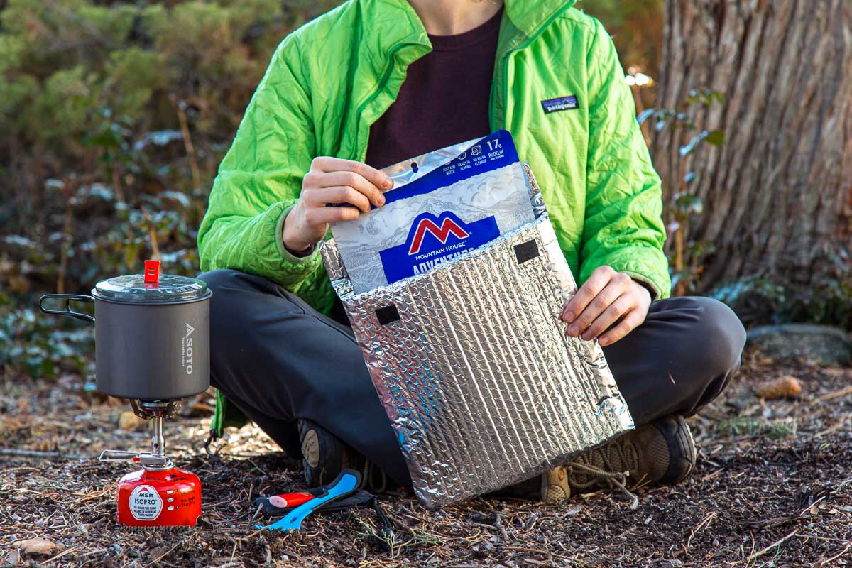 Megan is sitting on the ground placing a backpacking meal inside an insulated pouch cozy
