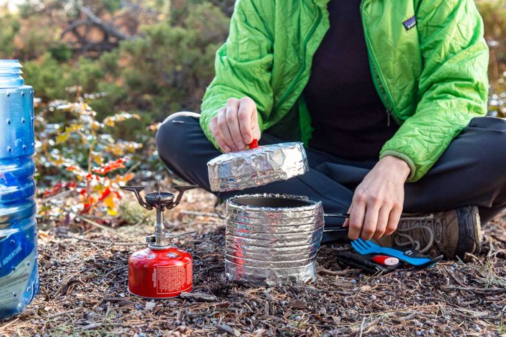 Megan is sitting on the ground outdoors placing the lid on a backpacking pot that is in a cozy