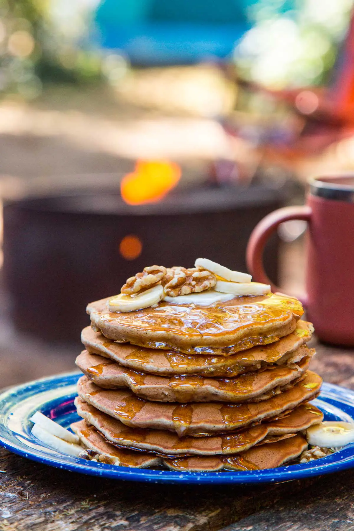 A stack of pancakes on a blue plate, topped with slices of bananas and walnuts and a camping scene in the background.