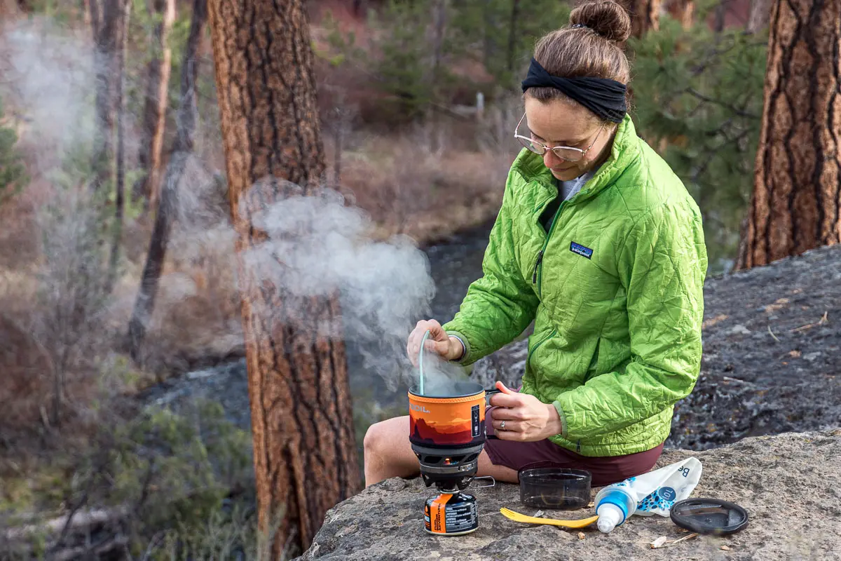 Megan is sitting on a rock cooking over a Jetboil backpacking stove 