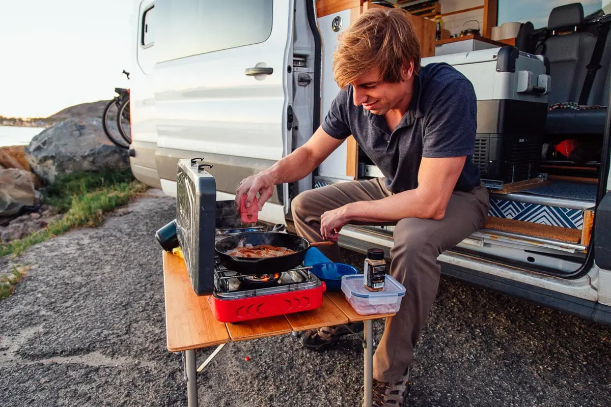 Michael sits on the stoop of a campervan and cooks at a campstove.