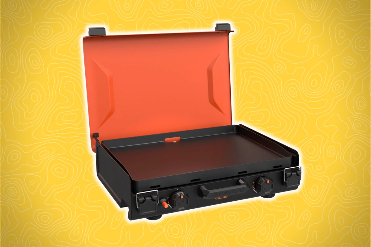 A portable black griddle with an orange lid sits open, showing a flat surface and two knobs on the front against a yellow background.