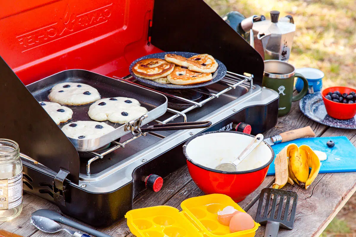 Pancakes on a griddle over a camp stove with various camp cooking utensils in frame