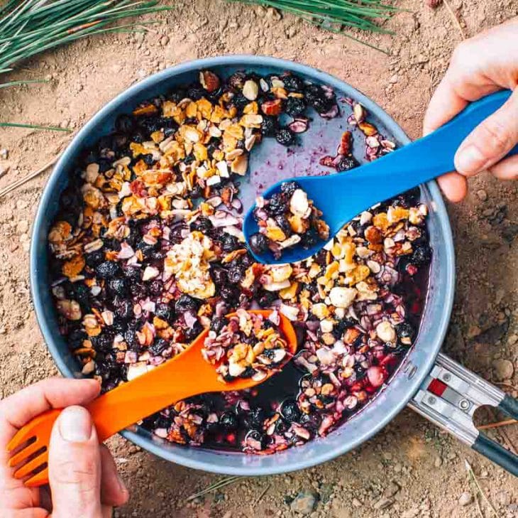 Blueberries topped with granola in a skillet with two hands and spoons reaching in
