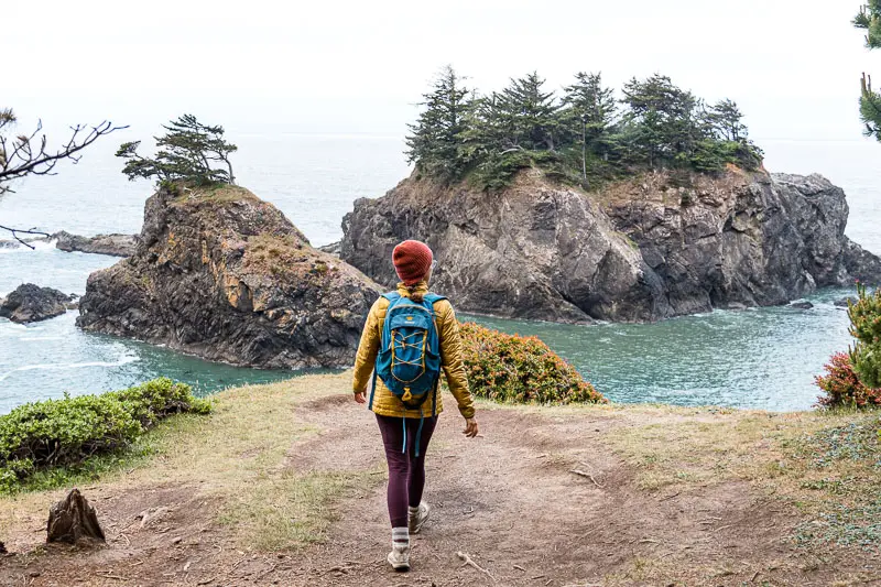 Megan walks on a dirt path towards the edge of a bluff with sea stacks in the distance.